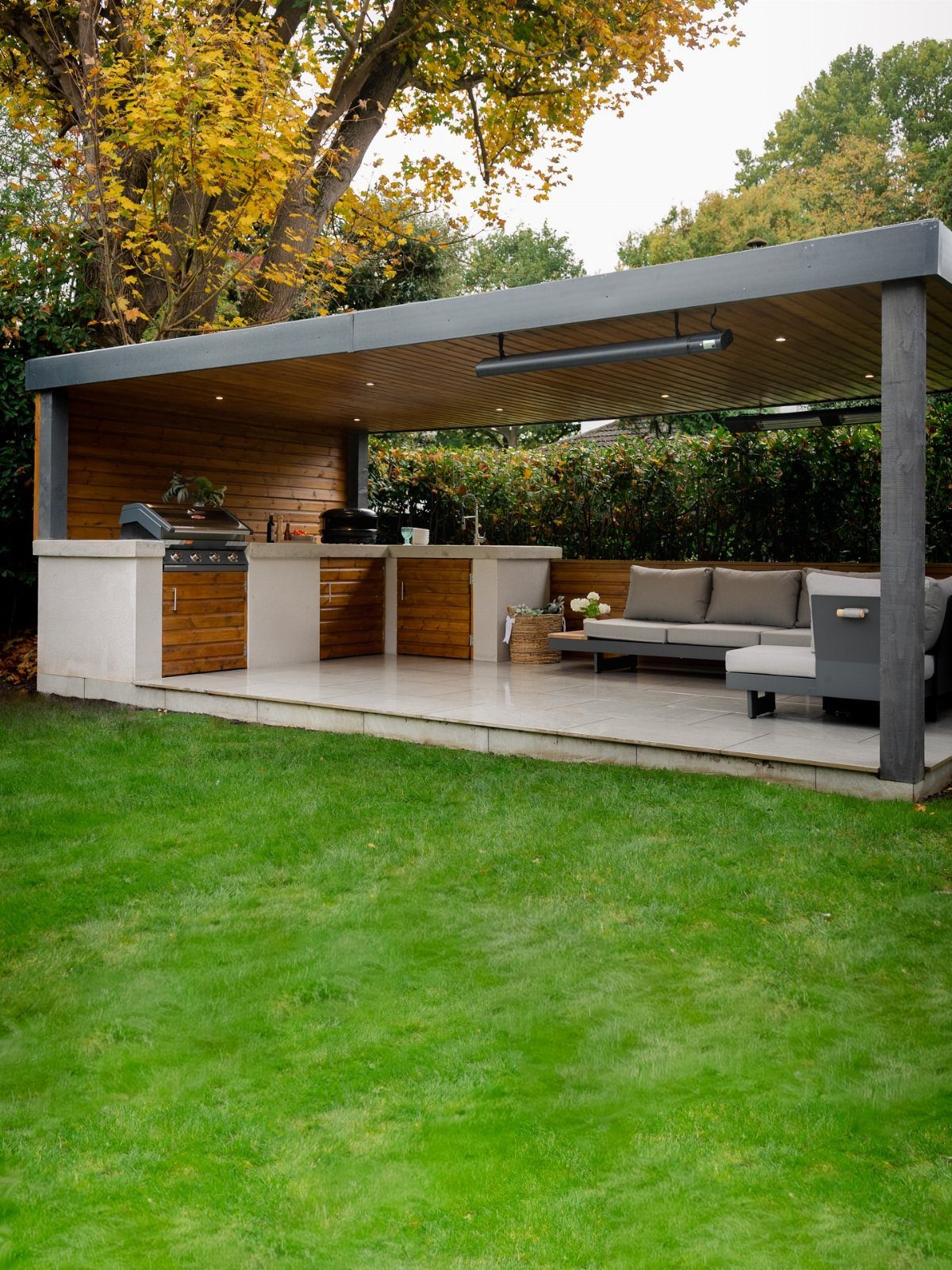 A modern outdoor kitchen with wooden cabinets and a grey sofa under a dark pergola.