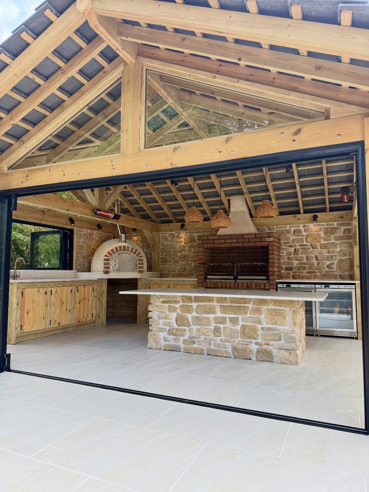An outdoor kitchen with stone walls, a brick grill, and a pizza oven under a wooden pavilion.