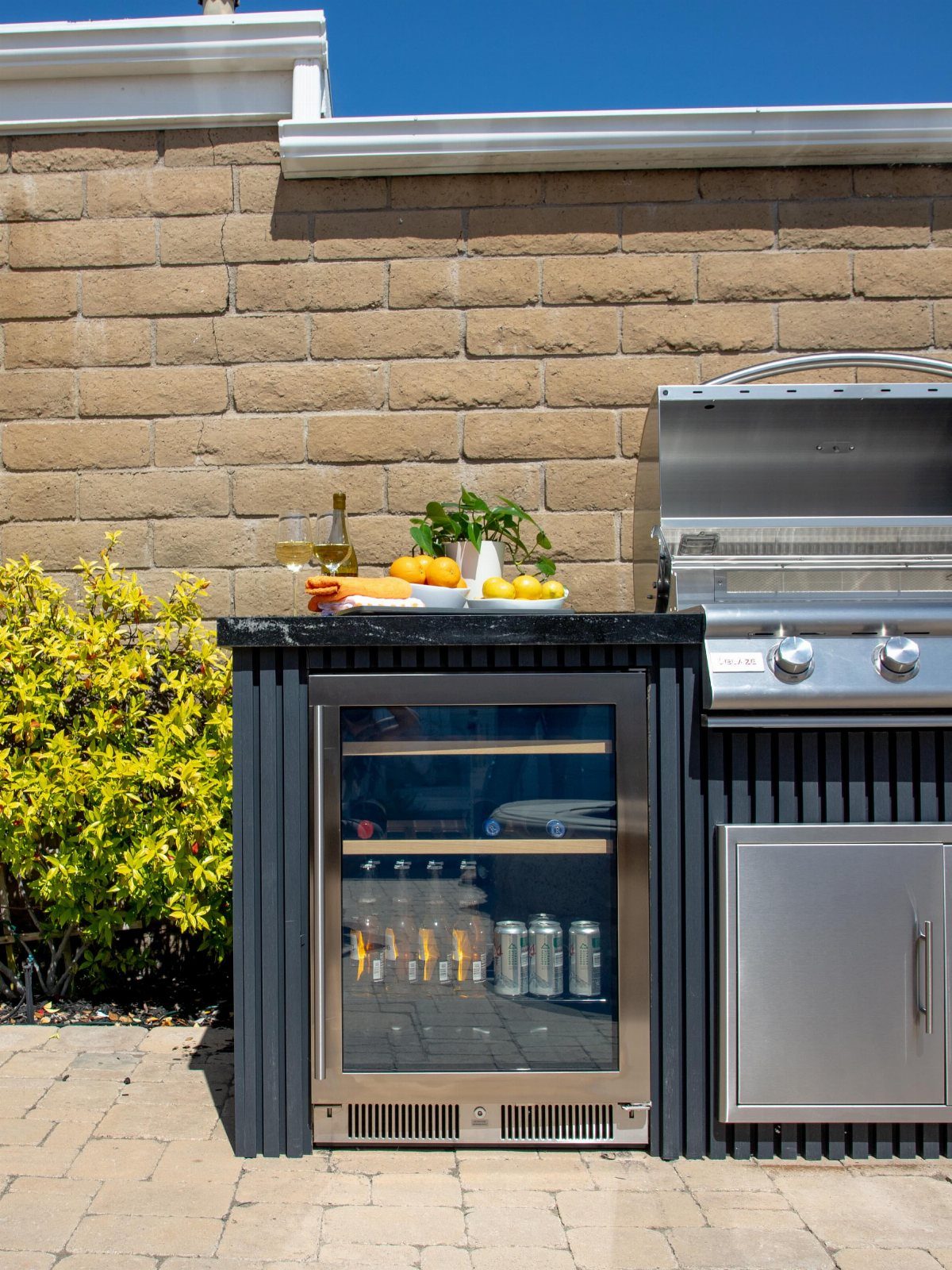 A stainless steel bar fridge with a glass door sits under a black outdoor kitchen countertop.