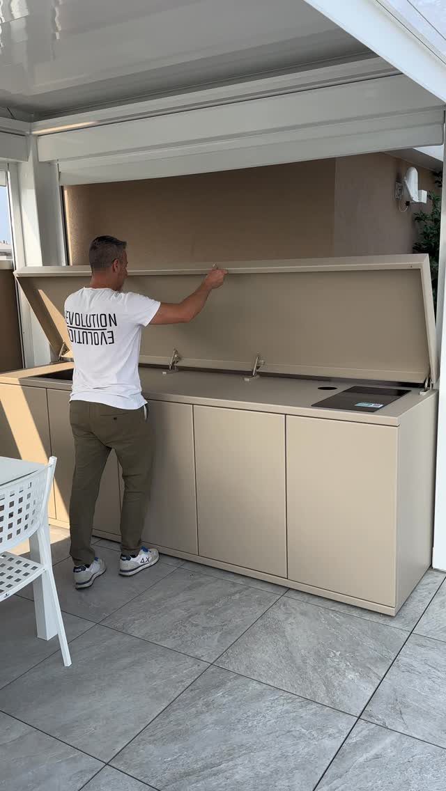 A man opening the lid of a modern beige outdoor kitchen unit on a patio.