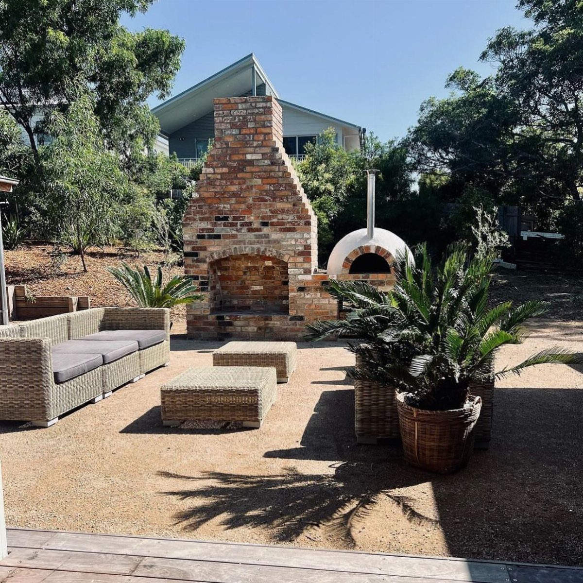 Brick outdoor fireplace and white pizza oven on a gravel patio with wicker lounge furniture.