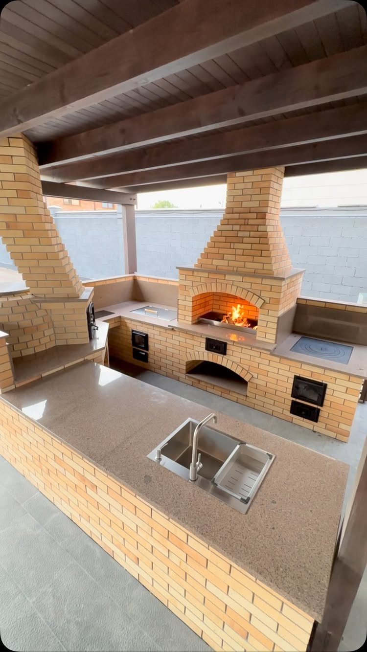 A brick outdoor kitchen with a built-in pizza oven and sink under a wooden pergola.