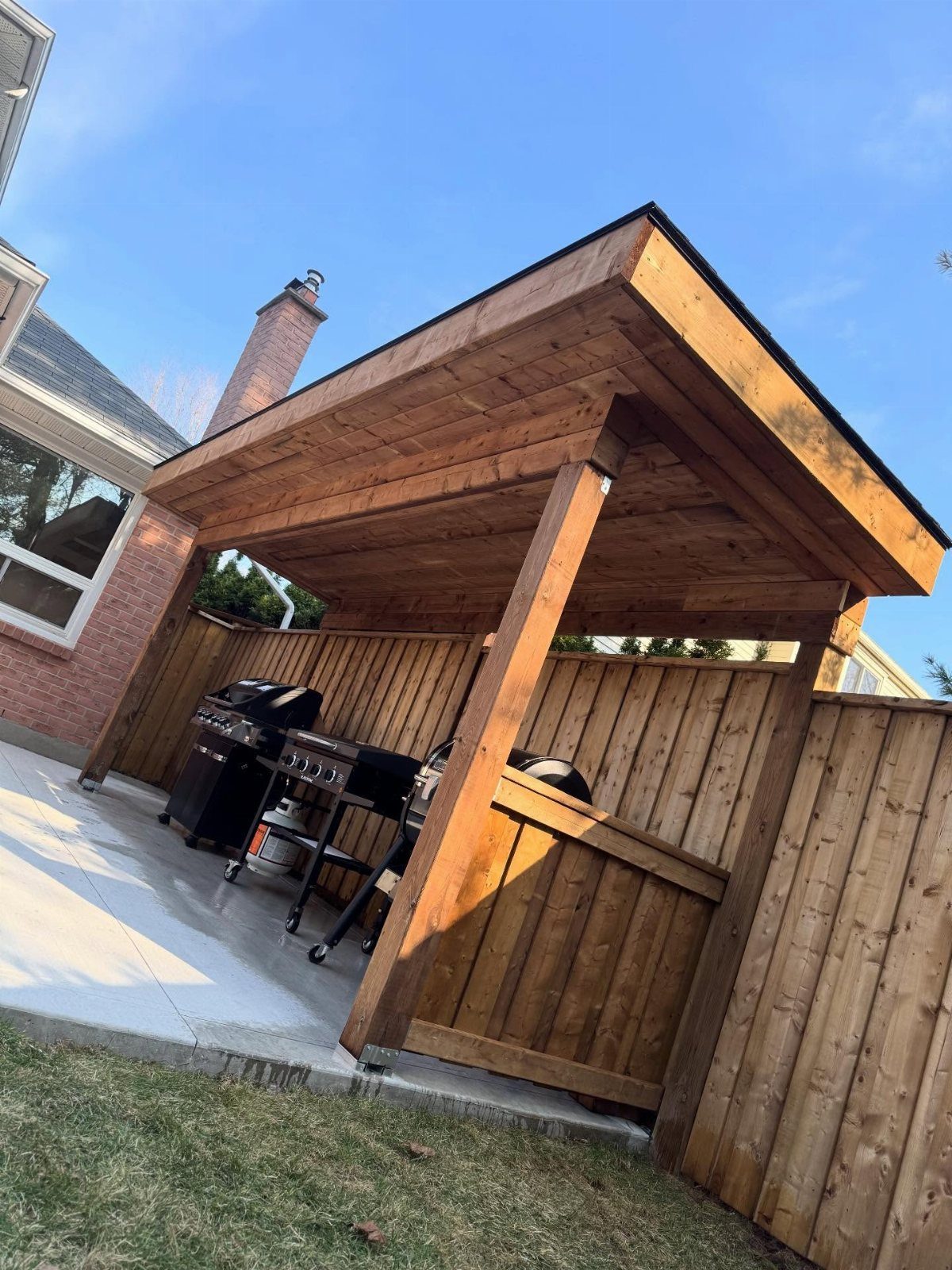A wooden lean-to shelter covering a gas grill on a concrete patio next to a fence.