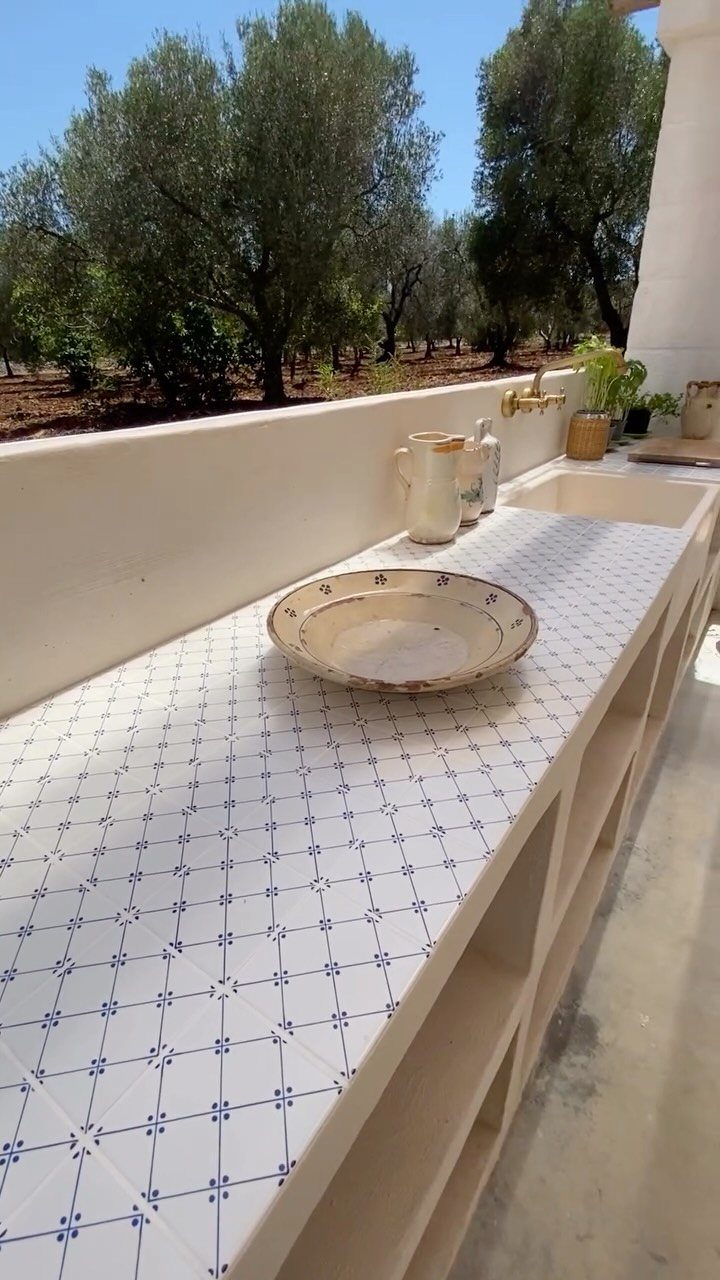 White tiled outdoor countertop with blue patterns, featuring built-in shelving and a sink overlooking olive trees.