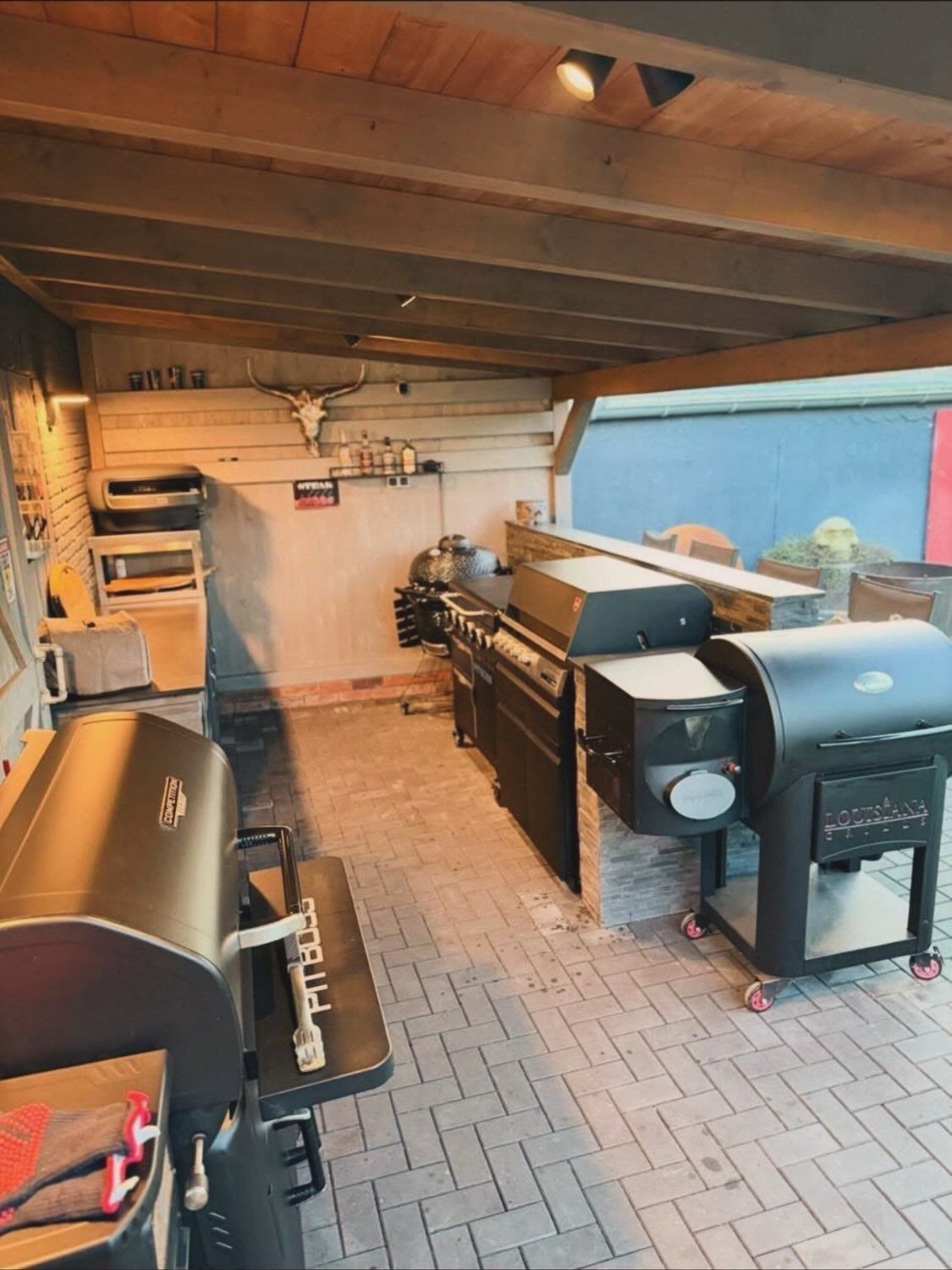 Several black outdoor grills and a smoker arranged under a wooden patio cover with ceiling lights.