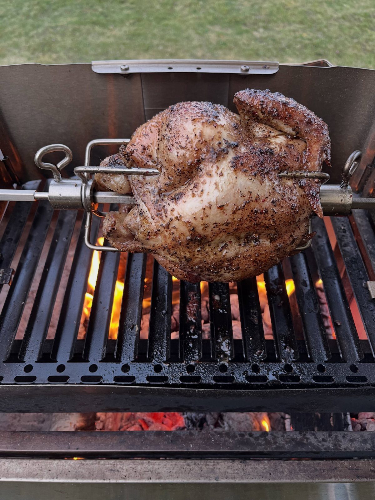 A seasoned whole chicken cooking on a rotisserie spit over hot glowing charcoal flames.