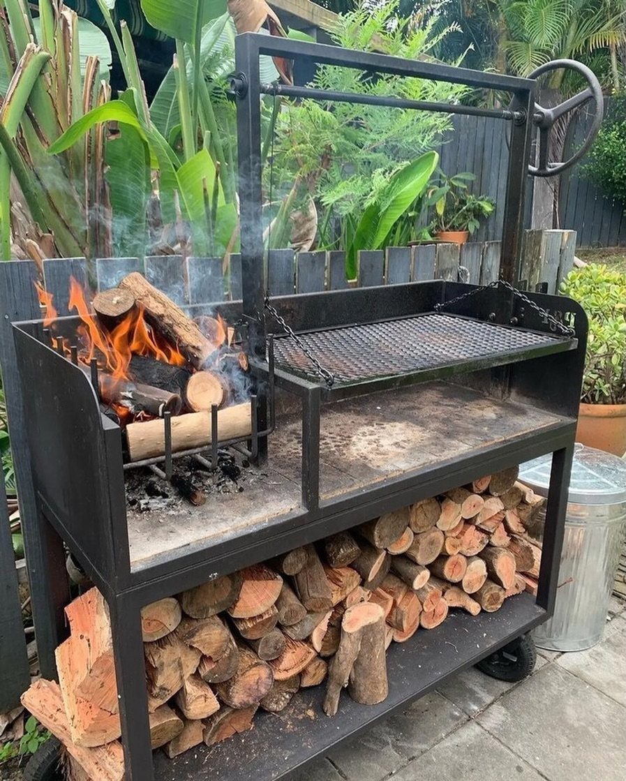 Black metal outdoor grill station with burning logs and stacked firewood underneath.
