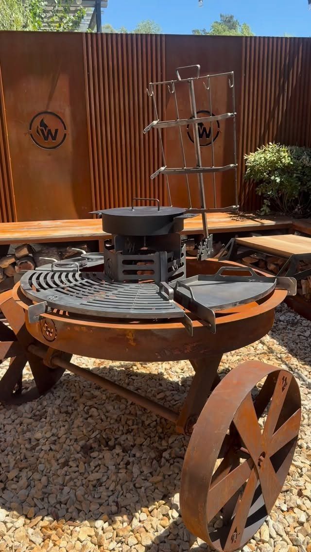 A large, rusted metal outdoor grill station with wheels sitting on a gravel surface.