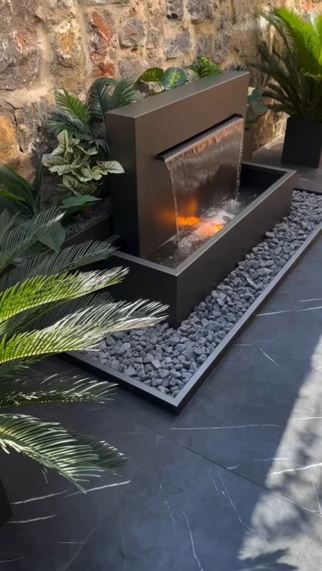 A modern dark grey rectangular water fountain flowing into a stone-filled basin surrounded by green plants.