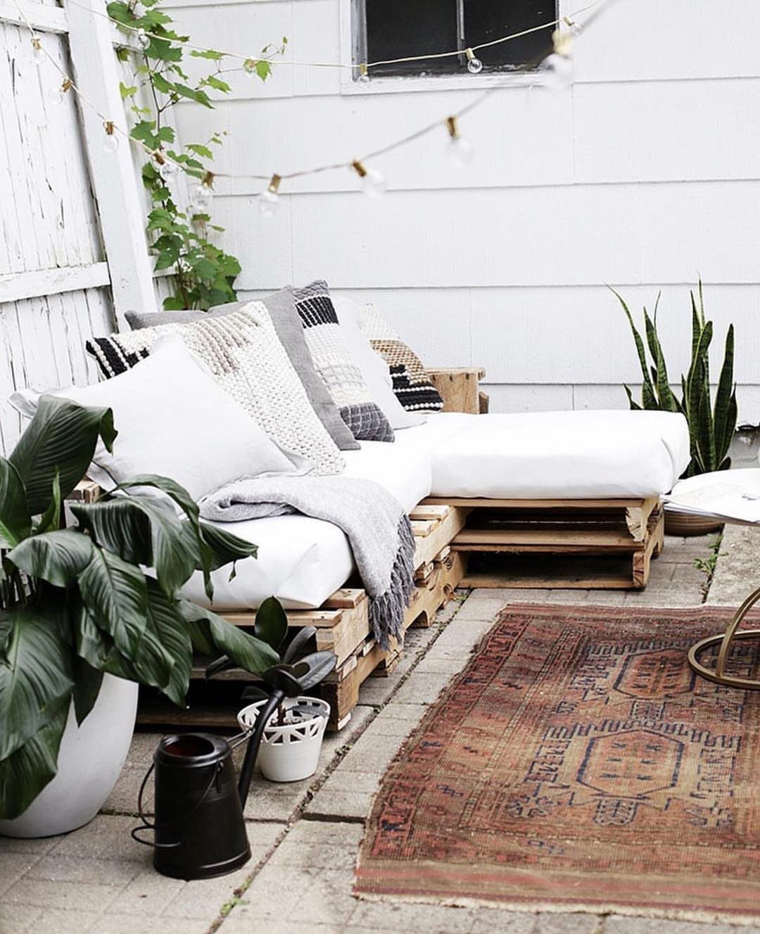 White pallet seating with various decorative pillows, a grey throw, and a patterned rug on a patio.
