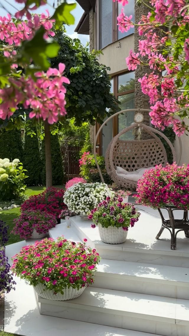 Colorful flowering plants in white baskets surround a hanging egg chair on a white stone patio.