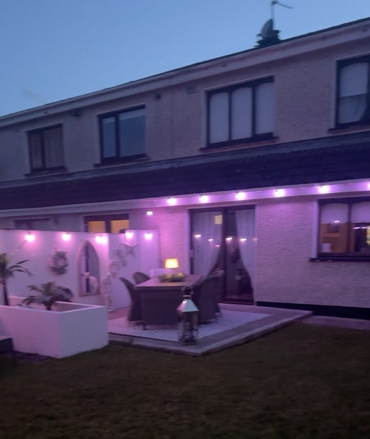 A patio dining set illuminated by pink string lights along a white garden wall at dusk.
