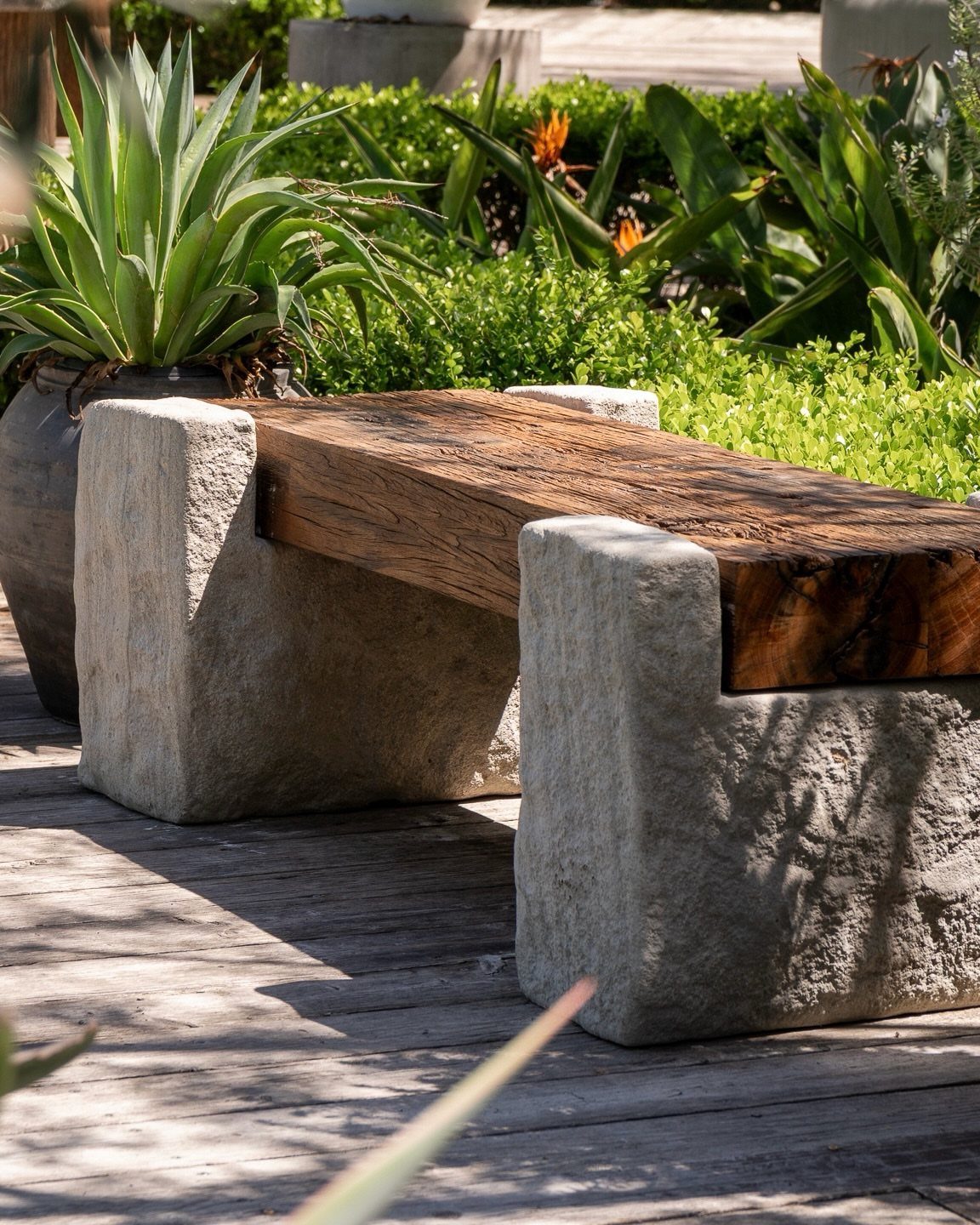 A weathered wood plank bench with stone supports sits on a wooden deck among green plants.