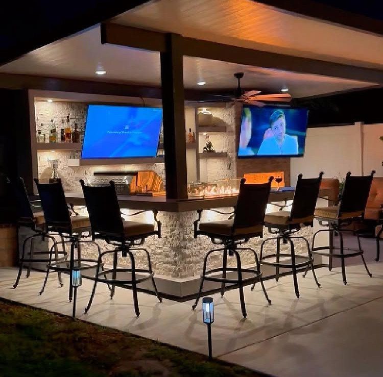 Modern outdoor bar with stone counter, black stools, and two televisions under a covered roof structure.