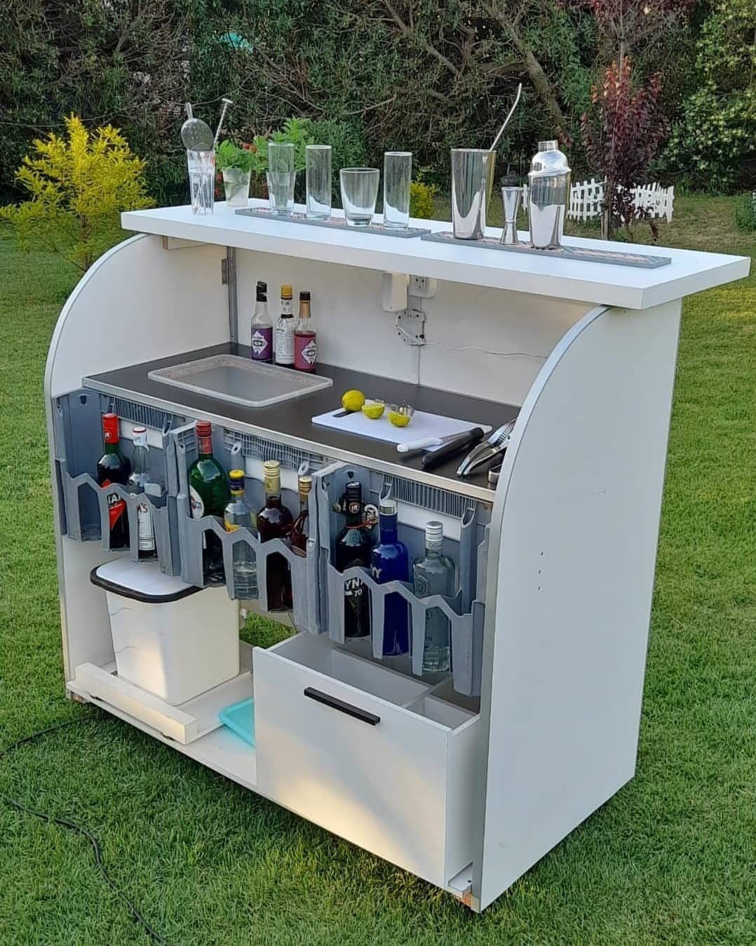 A white portable outdoor bartending station stocked with liquor bottles and glassware on a green lawn.