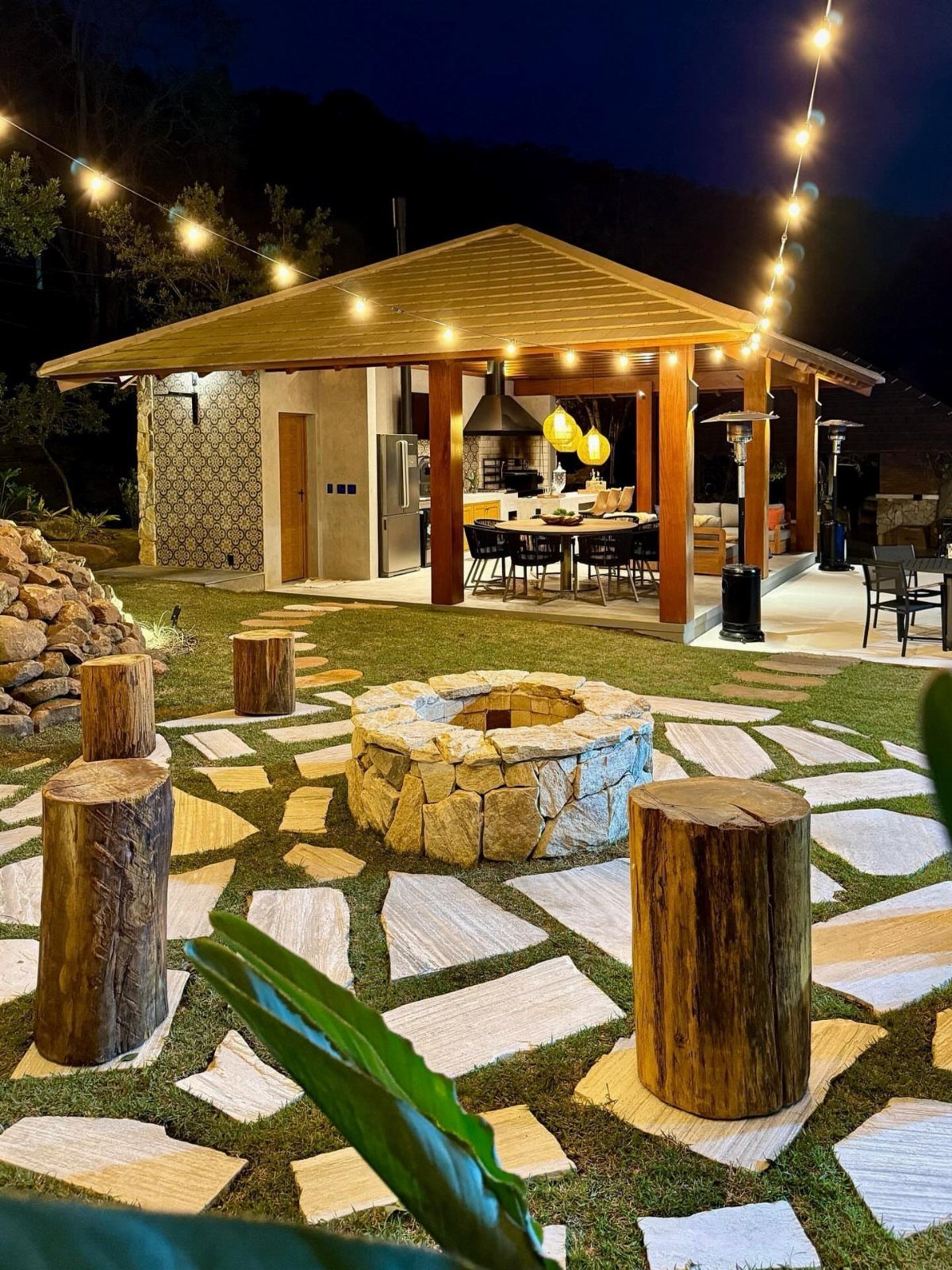 Outdoor patio at night with string lights, a stone fire pit, and a wooden kitchen pavilion.
