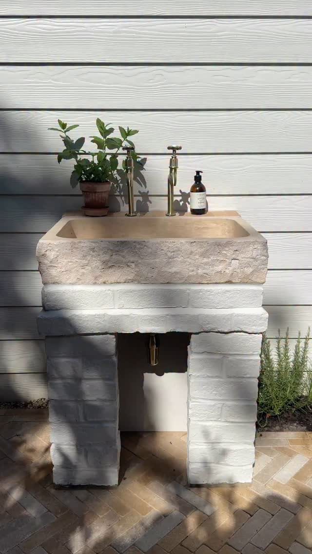 A stone outdoor sink with brass faucets sits on a white brick pedestal against siding.