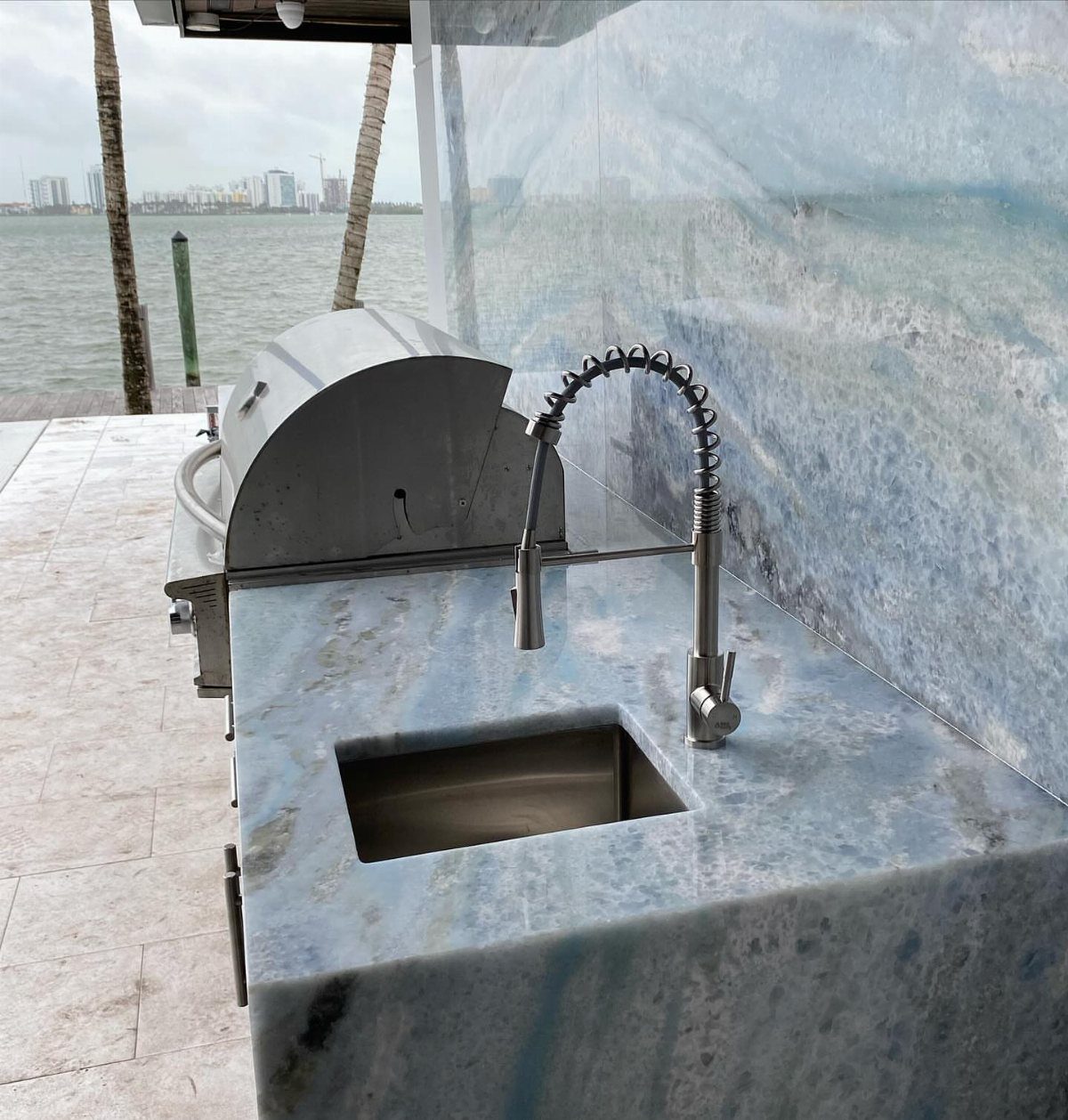Stainless steel grill and sink built into a blue marble outdoor kitchen countertop by the water.