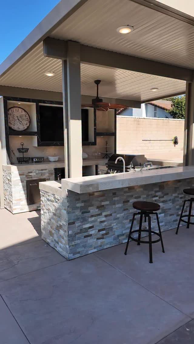 Stone-tiled outdoor kitchen island with a built-in grill, sink, and bar stools under a covered patio.