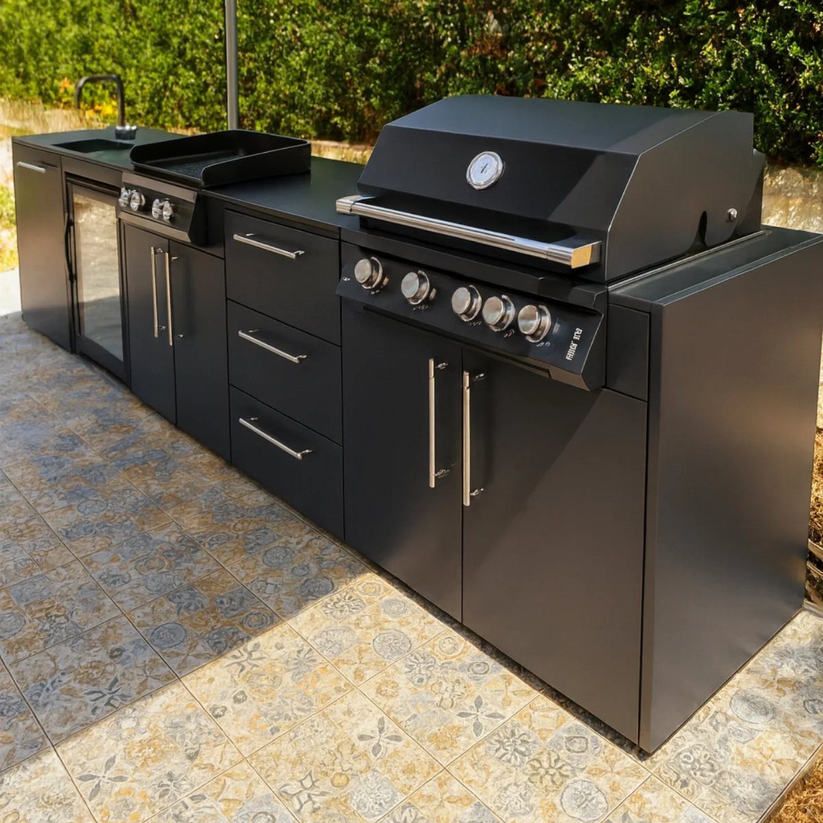 A black outdoor kitchen unit with a grill, griddle, sink, and cabinets on a tiled patio.