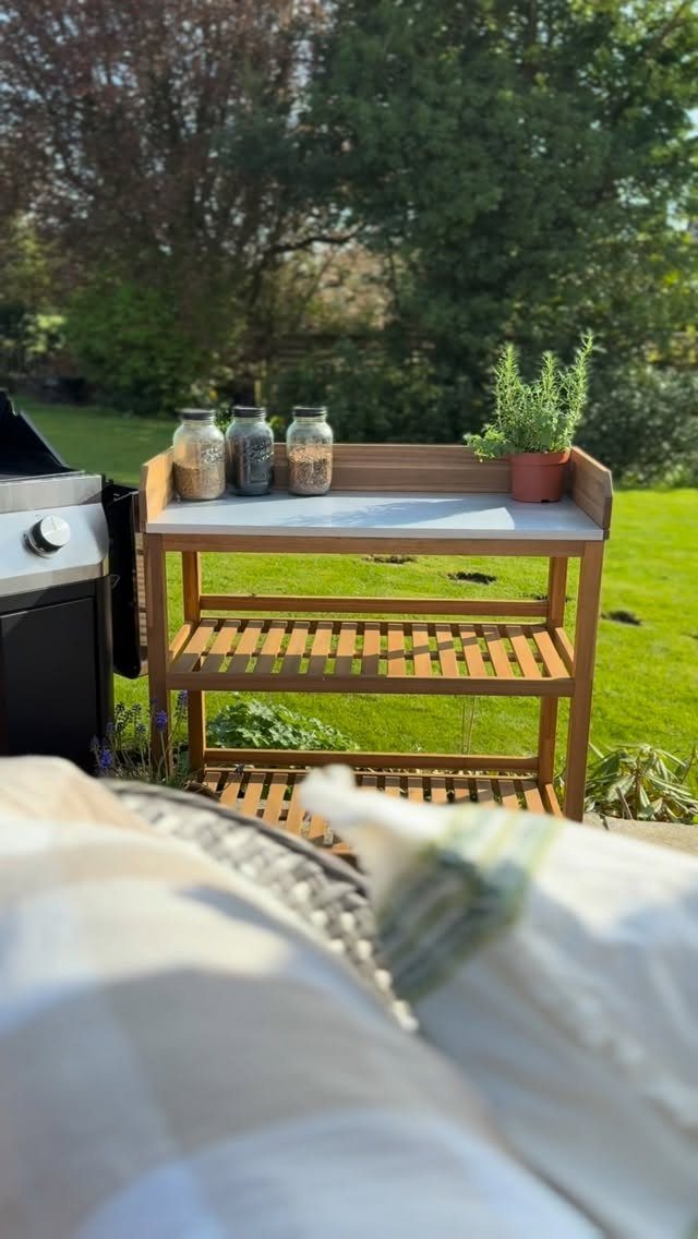 Wooden outdoor serving cart with glass jars and a potted plant on a green lawn.
