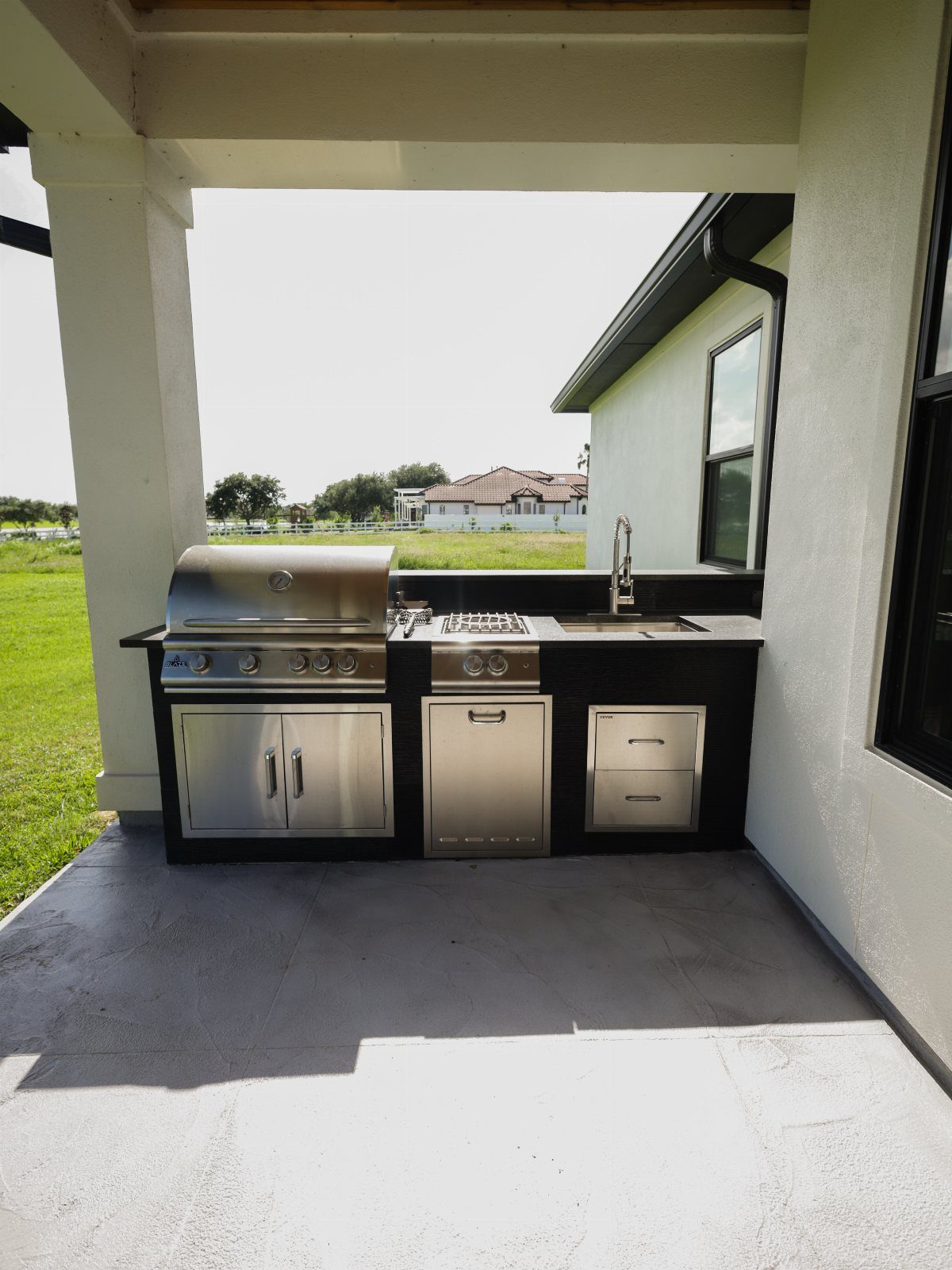 Stainless steel built-in outdoor kitchen with a large grill, sink, and cabinets under a patio.