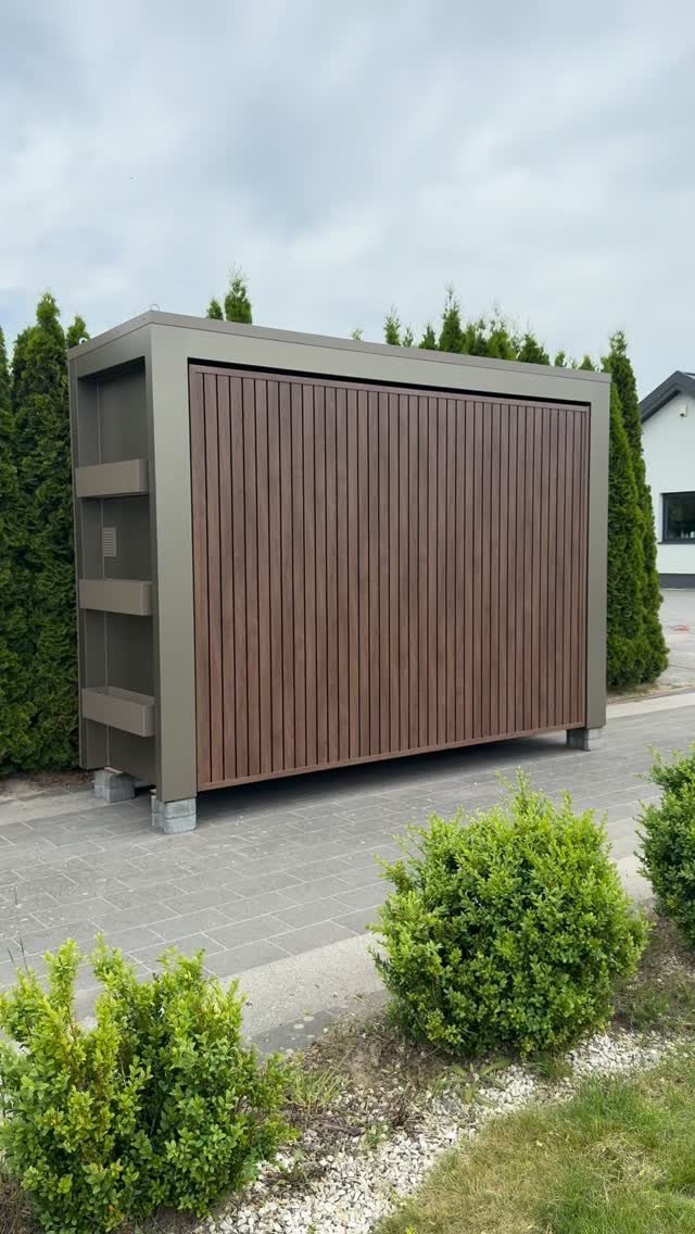 A large brown wooden storage shed with vertical slats positioned next to a tall green hedge.