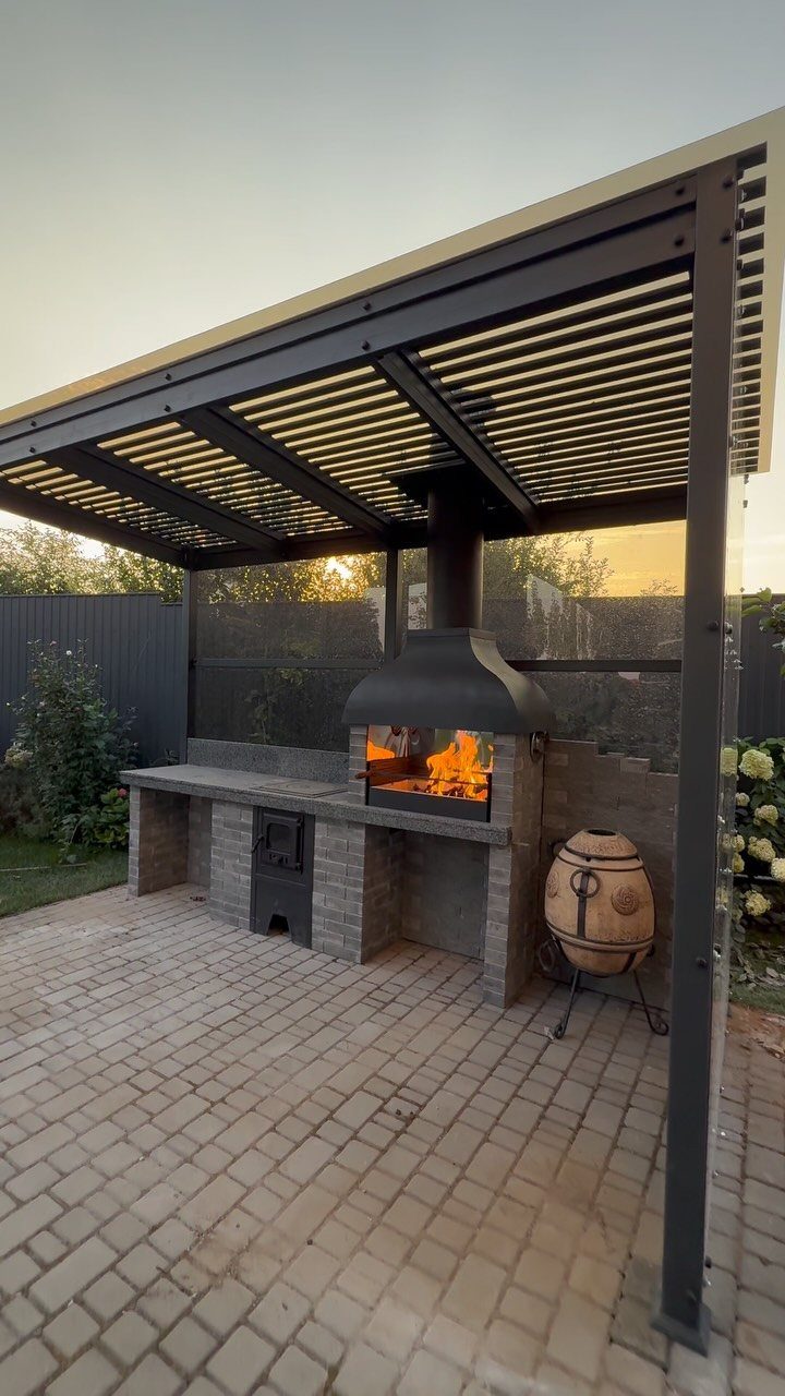 Built-in brick outdoor kitchen with a roaring fire under a dark metal pergola.