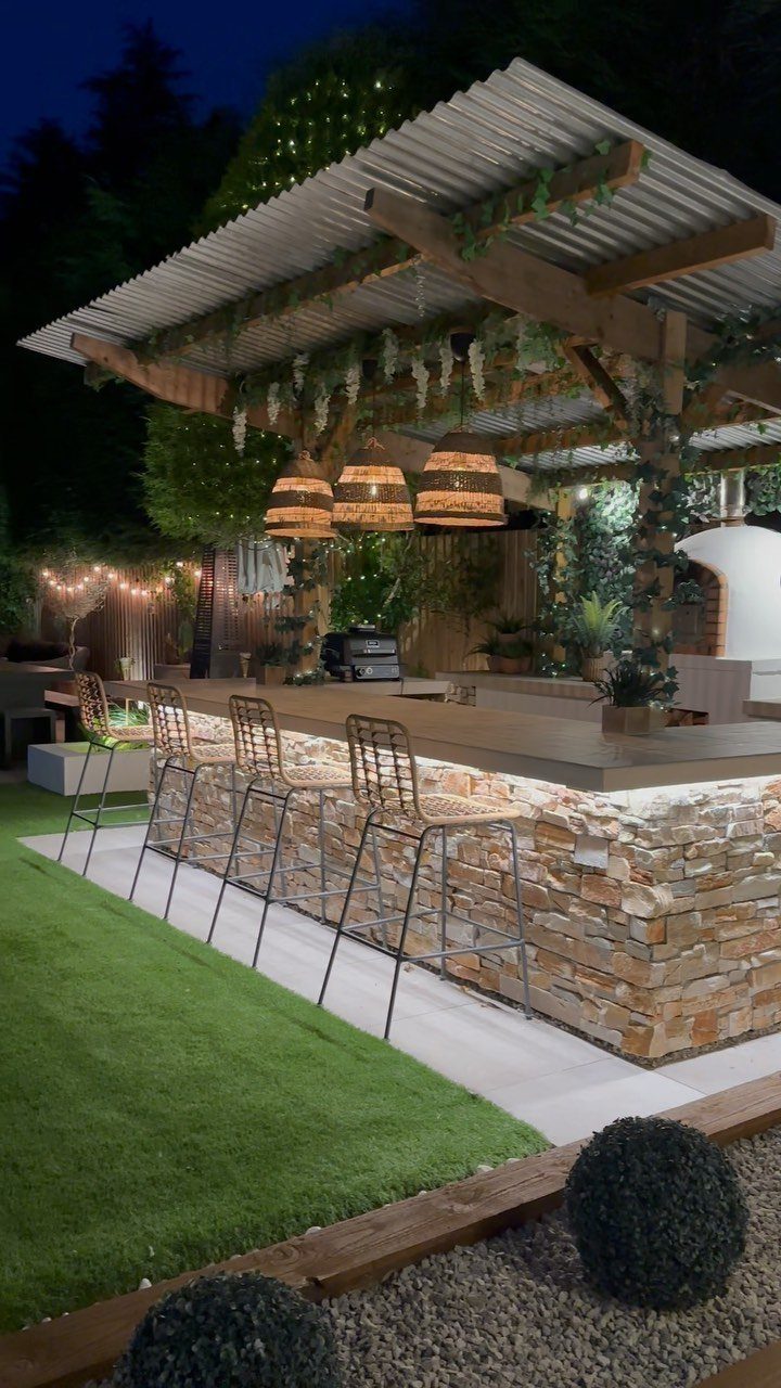 A lit outdoor kitchen bar with stone cladding and a wooden pergola structure at night.