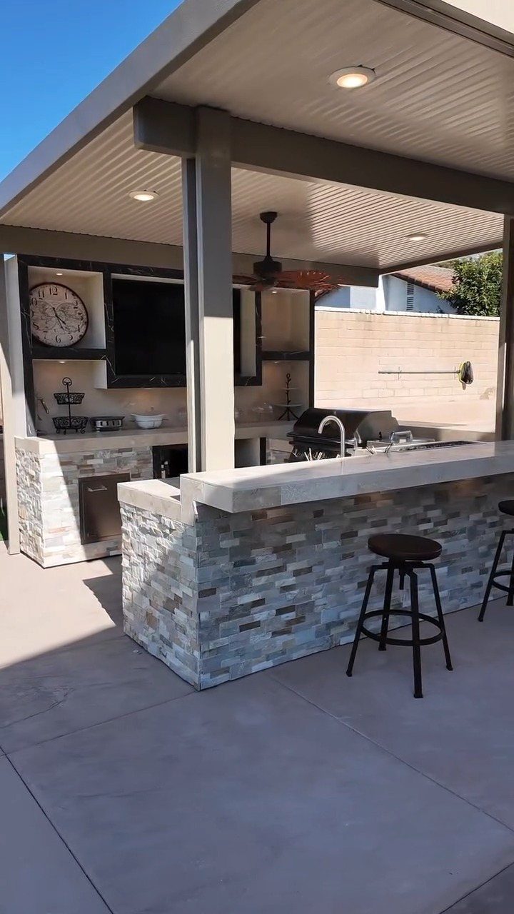 A modern outdoor kitchen with a stone island, built-in grill, and seating under a covered patio structure.