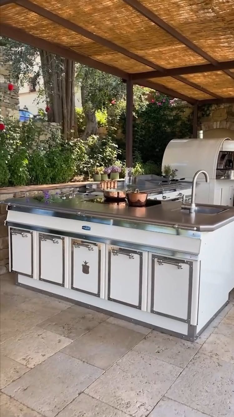 White outdoor kitchen island with stainless steel accents under a bamboo reed pergola on a stone patio.