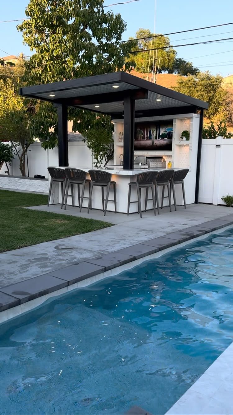 Modern outdoor kitchen with a black pergola, white counters, and bar stools next to a swimming pool.