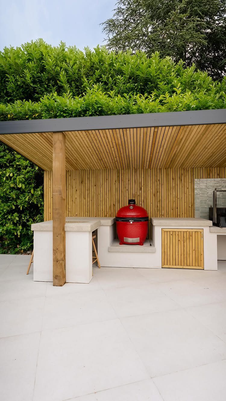 Red kamado grill set into a white stone outdoor kitchen under a wooden pergola.