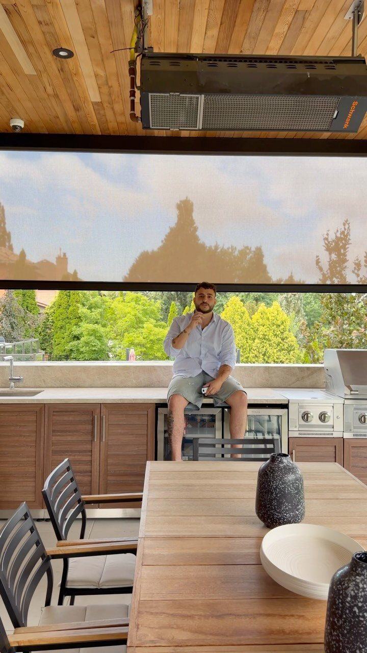 A man sits in a modern outdoor kitchen under a wooden ceiling with a retractable shade screen.