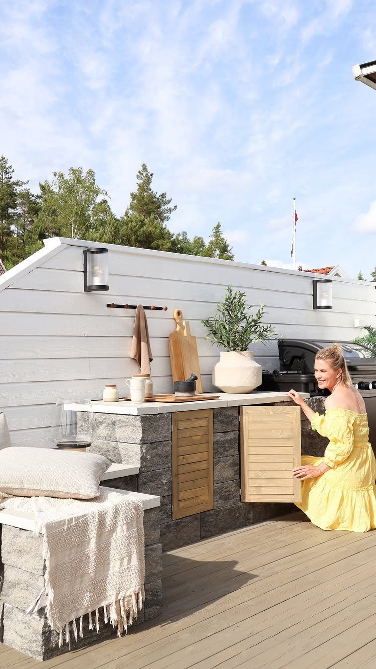 A woman kneels beside a stone outdoor kitchen with light wood cabinets on a wooden deck.