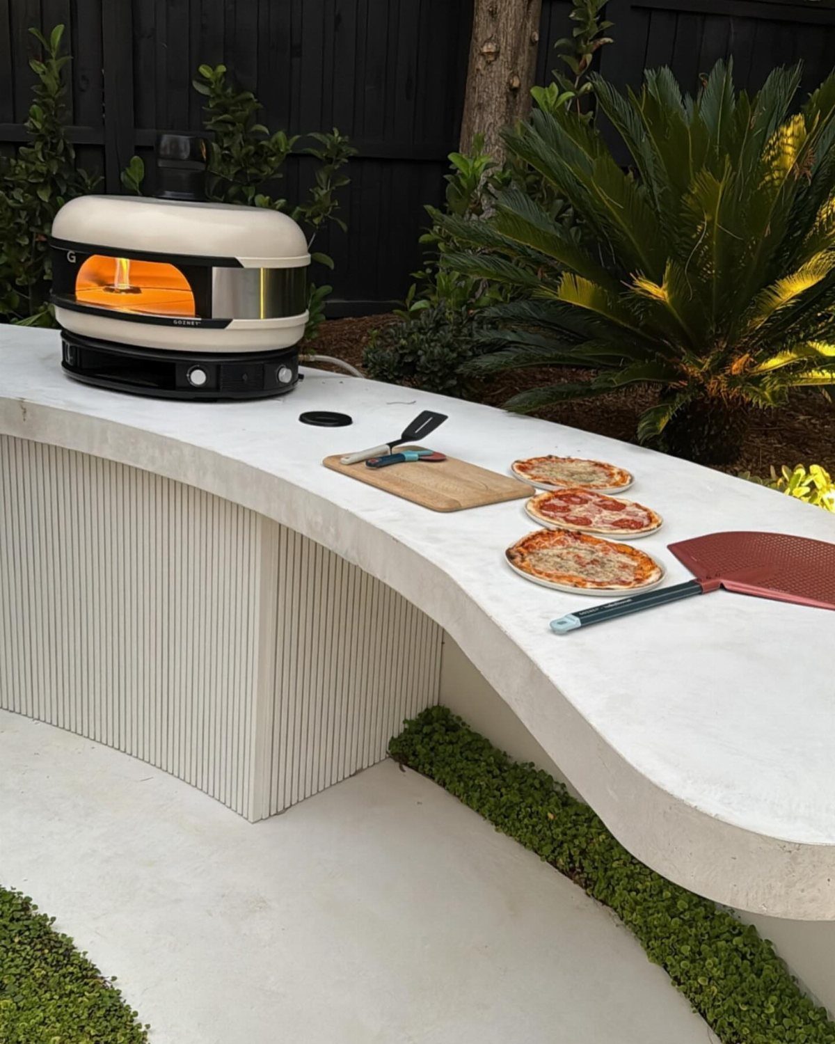 A white curved outdoor counter with a portable pizza oven and three cooked pizzas.