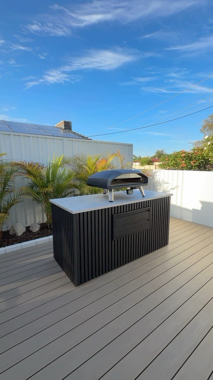 A black outdoor pizza oven sits on a white countertop of a modern outdoor kitchen island.
