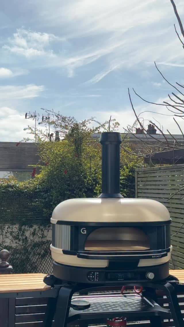 A cream-colored portable pizza oven sitting on a black stand on an outdoor wooden table.