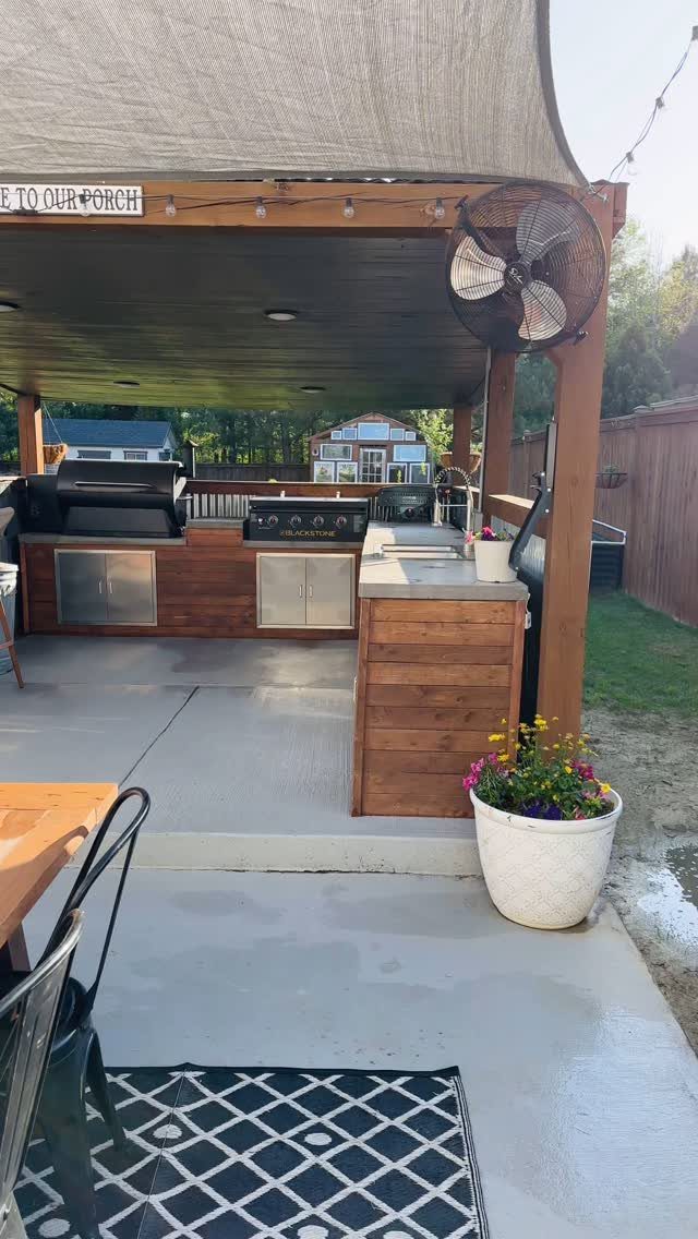 A wooden outdoor kitchen with a built-in grill and stainless steel cabinets under a covered patio.