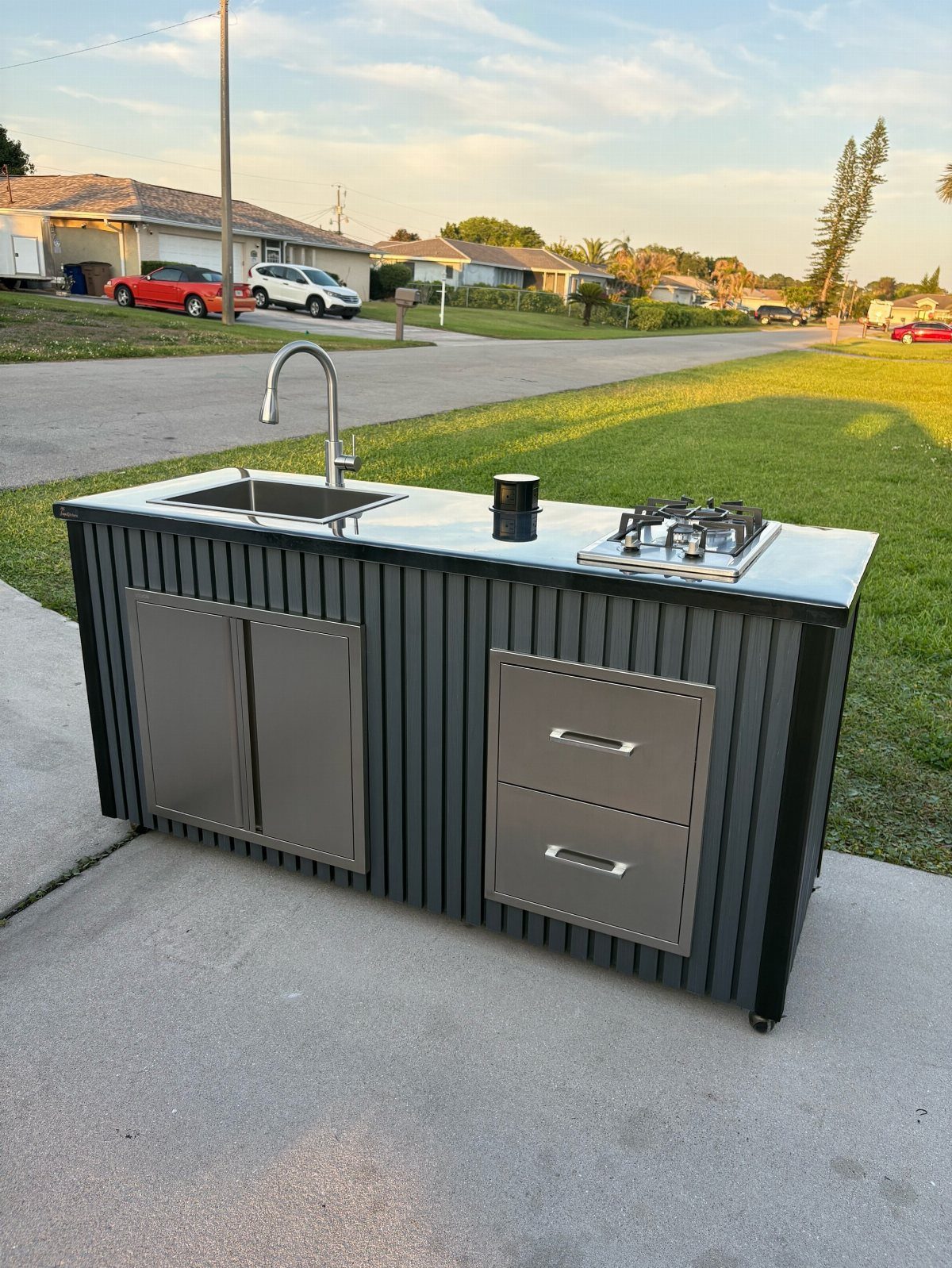 A black corrugated metal mobile outdoor kitchen cart with a sink and gas stove on a driveway.