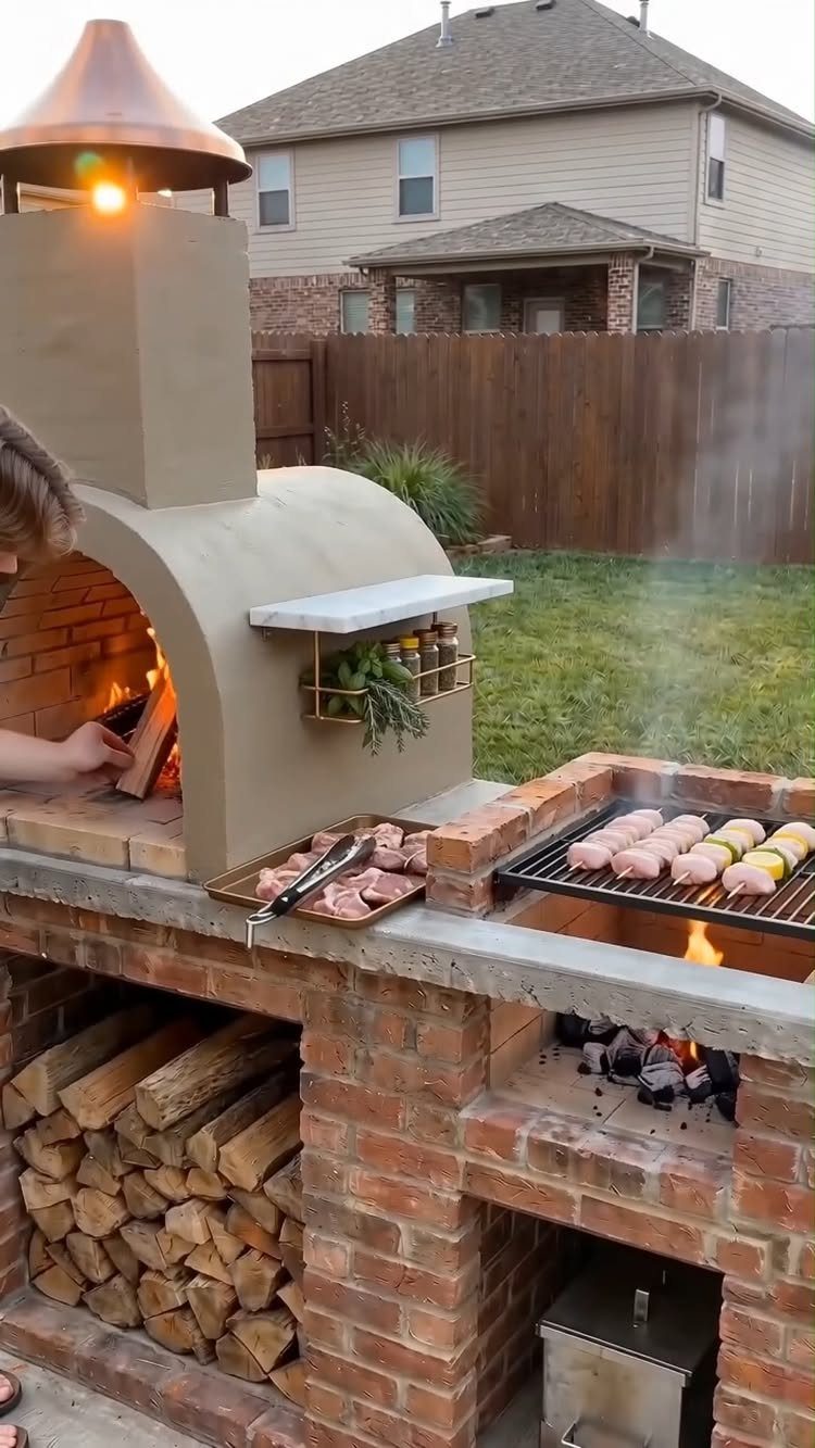 A brick outdoor kitchen with a pizza oven, grill, and stacked firewood in a backyard.