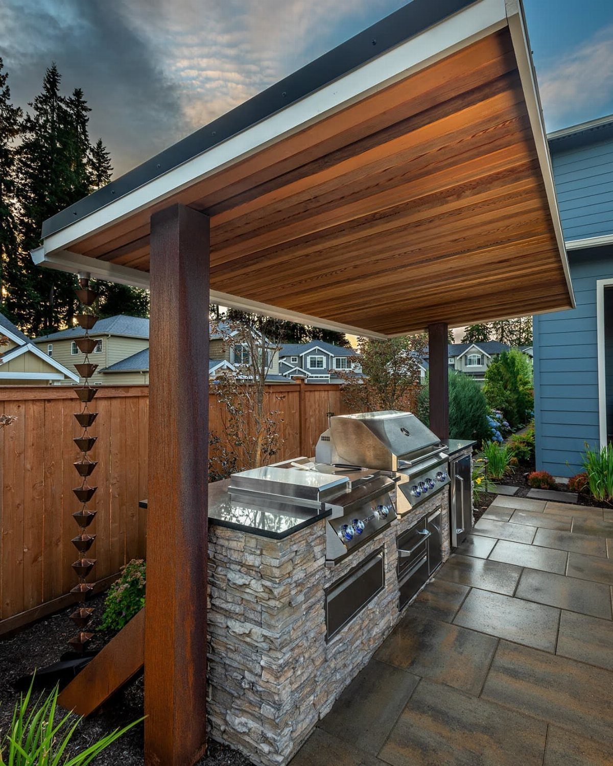 A stone outdoor kitchen with a stainless steel grill under a wooden roof structure.