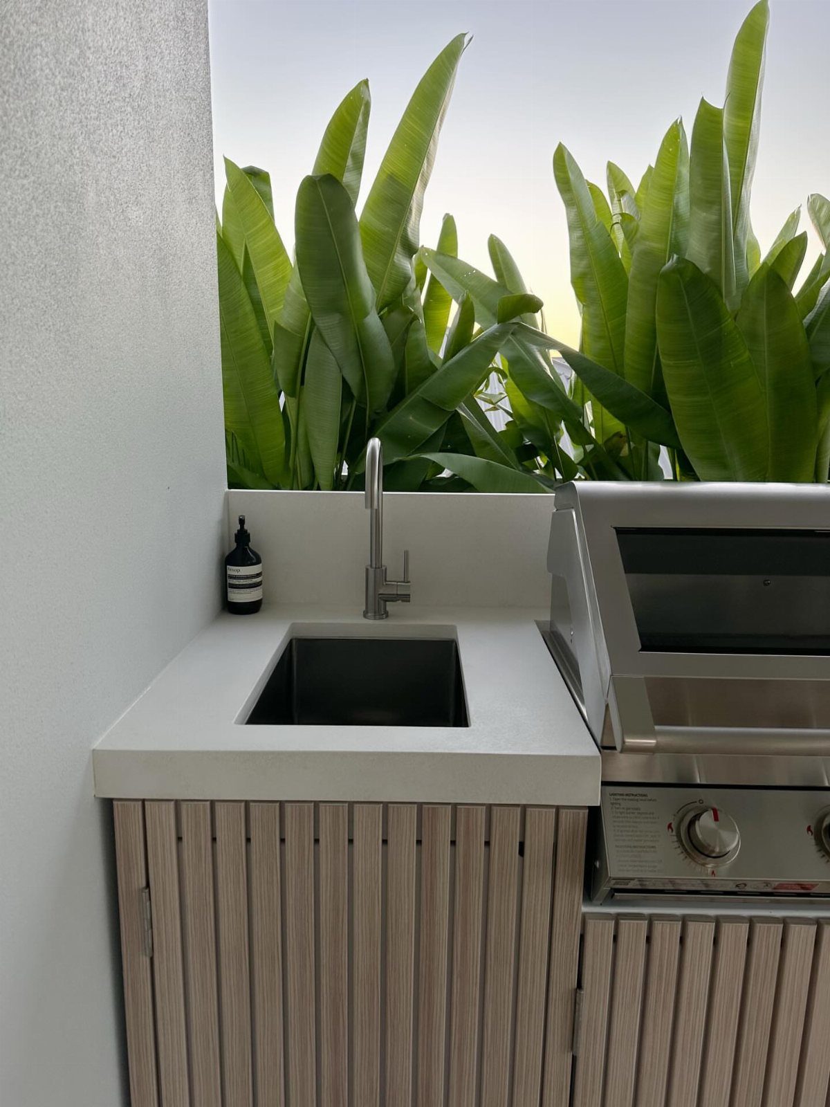 A modern outdoor kitchen with a white countertop, black sink, and stainless steel grill.