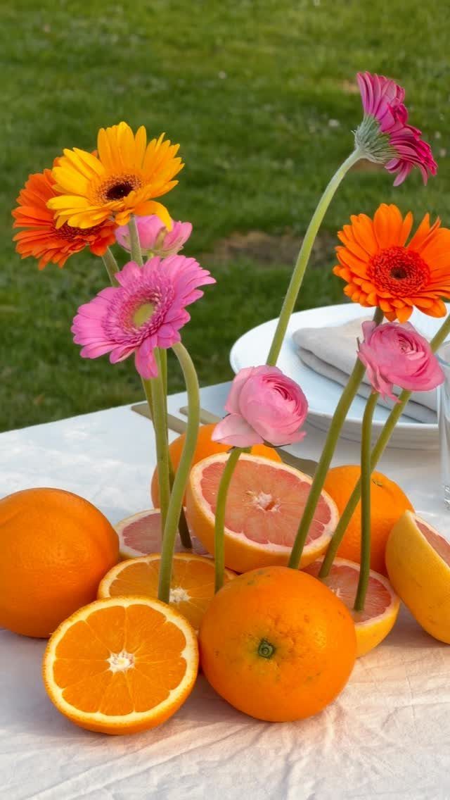 Colorful gerbera daisies and pink ranunculus flowers arranged with sliced citrus fruits on a white table.