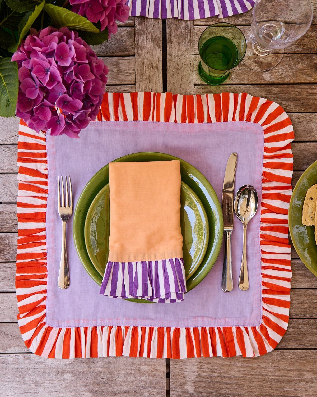 An orange napkin with purple stripes sits on a green plate atop a lavender placemat.