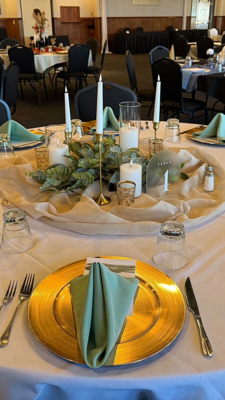 A decorated round dining table featuring gold chargers, green napkins, and white candles with eucalyptus greenery.