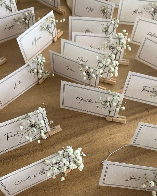 Handwritten name place cards decorated with small white flowers and wooden clips on a wooden table.
