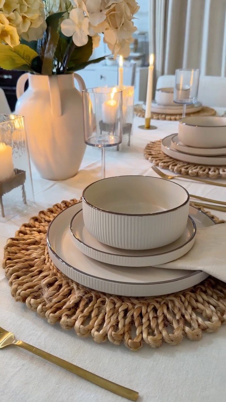 Stacked white ceramic plates and bowls on woven jute placemats with gold cutlery on a white tablecloth.