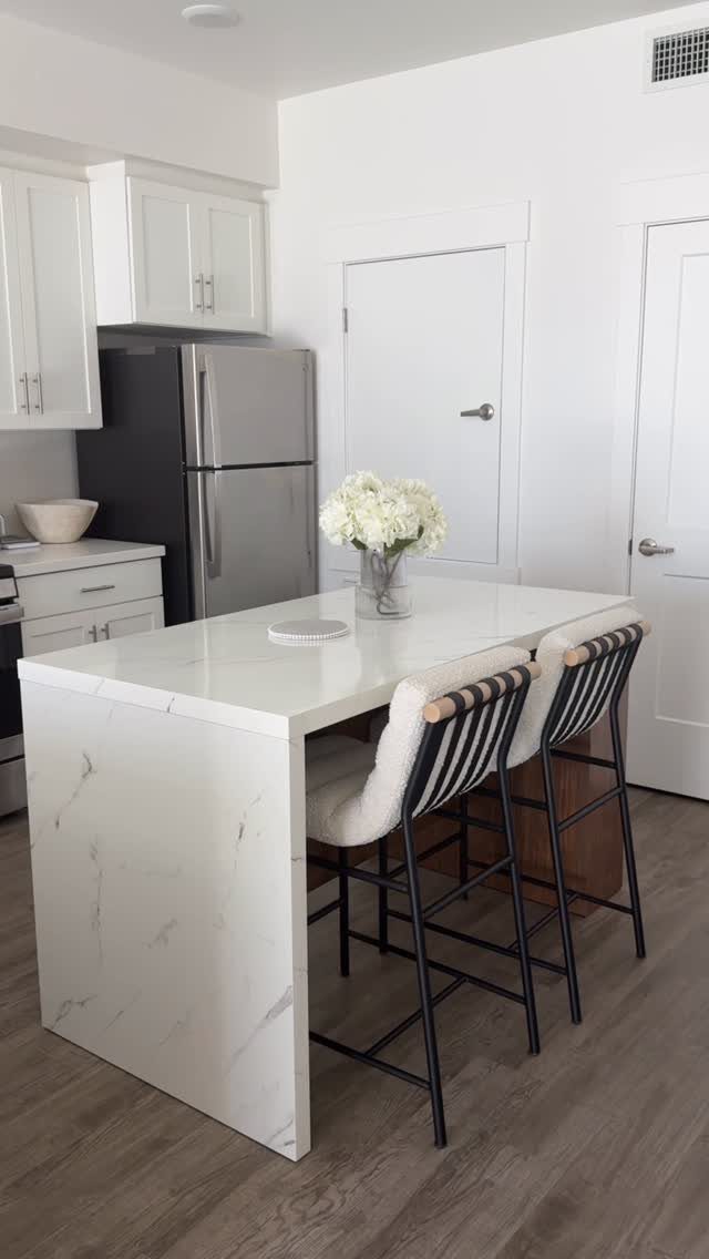 White marble kitchen island with two black bar stools and white flowers in a bright kitchen.