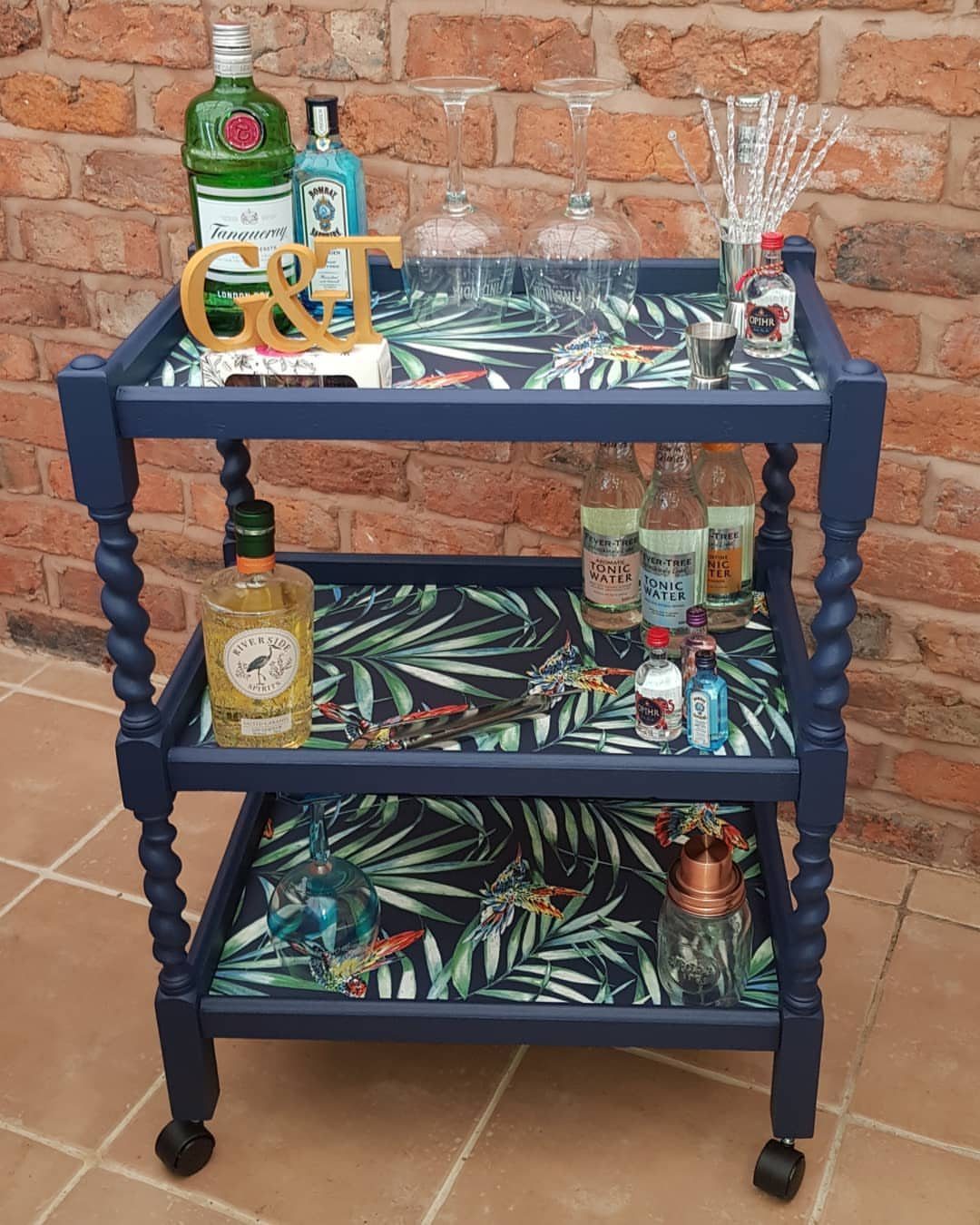 A navy blue three-tier wooden drinks trolley filled with various liquor bottles and glassware.